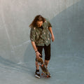 Photo of male, long-haired skateboarder standing with skateboard in skatepark bowl. He is wearing Blinky Bills Shirt No. 3, an olive coloured, over-sized, short-sleeve shirt with a print of different tattoo designs of vases.