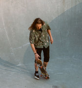 Photo of male, long-haired skateboarder standing with skateboard in skatepark bowl. He is wearing Blinky Bills Shirt No. 3, an olive coloured, over-sized, short-sleeve shirt with a print of different tattoo designs of vases.