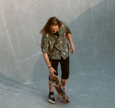 Photo of male, long-haired skateboarder standing with skateboard in skatepark bowl. He is wearing Blinky Bills Shirt No. 3, an olive coloured, over-sized, short-sleeve shirt with a print of different tattoo designs of vases.