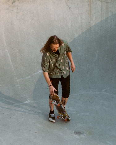 Photo of male, long-haired skateboarder standing with skateboard in skatepark bowl. He is wearing Blinky Bills Shirt No. 3, an olive coloured, over-sized, short-sleeve shirt with a print of different tattoo designs of vases.