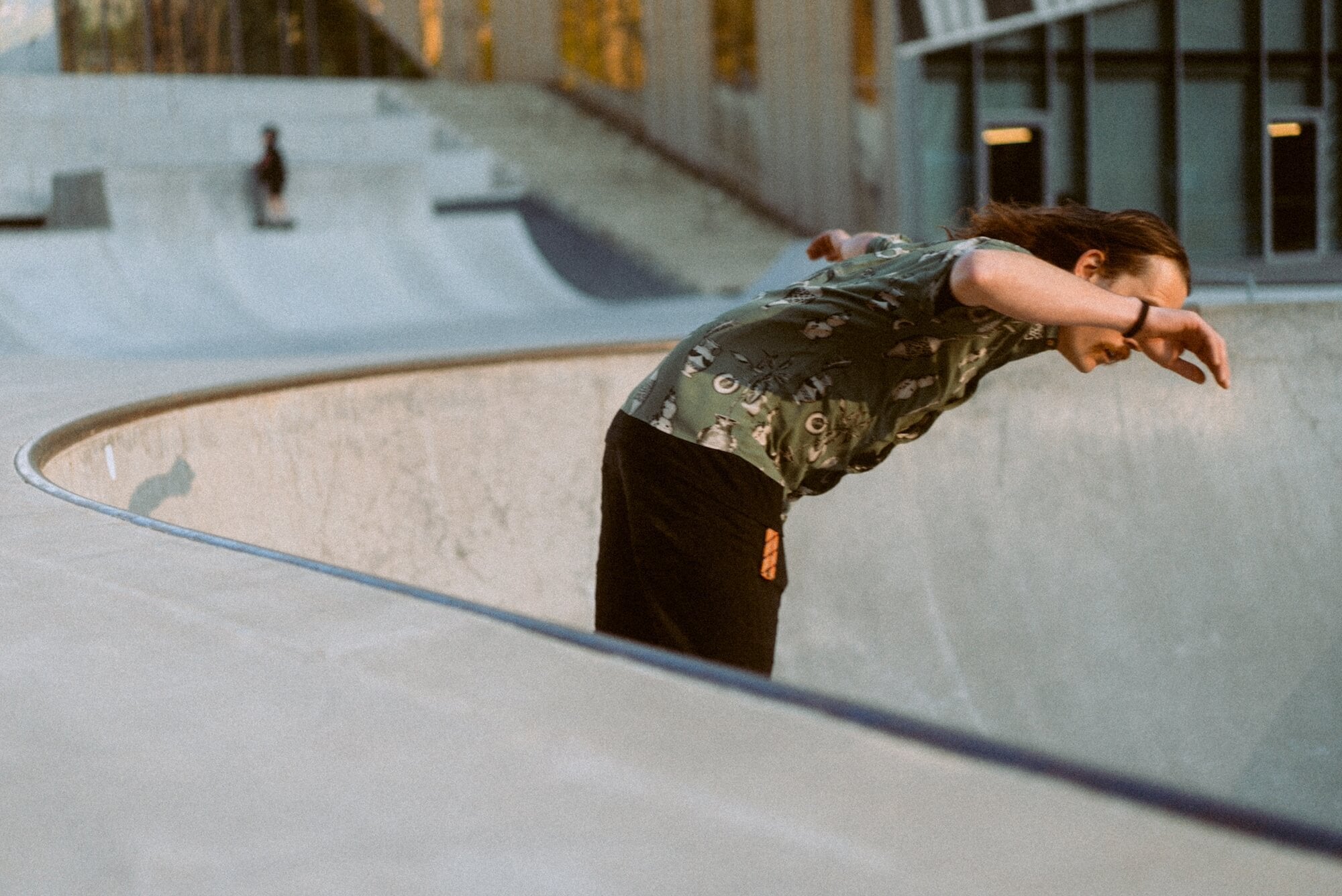 Photo of male, long-haired skateboarder skateboarding round a skatepark bowl. He is wearing Blinky Bills Shirt No. 3, an olive coloured, over-sized, short-sleeve shirt with a print of different tattoo designs of vases.