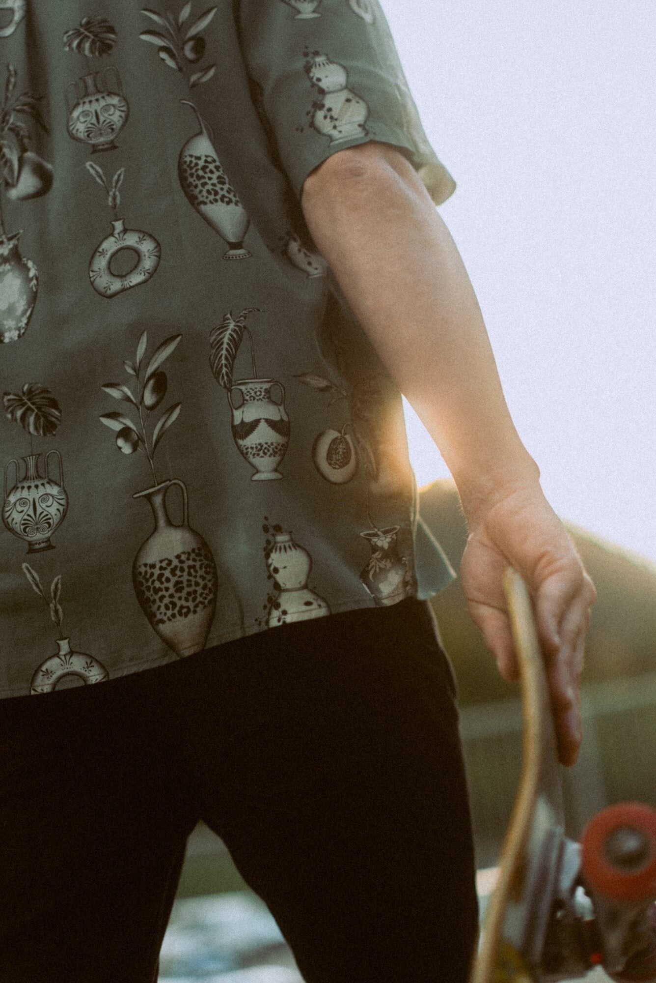 Close up photo of man holding a skateboard. He is wearing Blinky Bills Shirt No. 3, an olive coloured, over-sized, short-sleeve shirt with a print of different tattoo designs of vases.