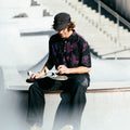 Photo of man with cap on sitting on skatepark bowl adjusting his skateboard wheels. He is wearing Blinky Bills Shirt No. 1, a black over-sized, short-sleeved shirt with a print of pink and purple tattoo-designed tigers.