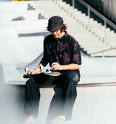 Photo of man with cap on sitting on skatepark bowl adjusting his skateboard wheels. He is wearing Blinky Bills Shirt No. 1, a black over-sized, short-sleeved shirt with a print of pink and purple tattoo-designed tigers.