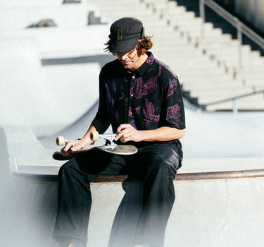 Photo of man with cap on sitting on skatepark bowl adjusting his skateboard wheels. He is wearing Blinky Bills Shirt No. 1, a black over-sized, short-sleeved shirt with a print of pink and purple tattoo-designed tigers.