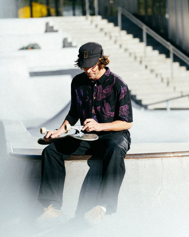 Photo of man with cap on sitting on skatepark bowl adjusting his skateboard wheels. He is wearing Blinky Bills Shirt No. 1, a black over-sized, short-sleeved shirt with a print of pink and purple tattoo-designed tigers.