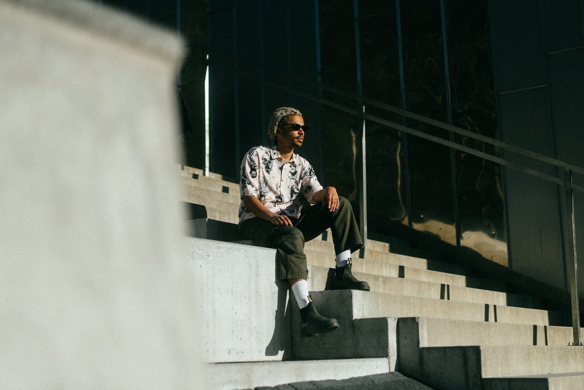 Photo of braided-haired man sitting on steps. He is wearing Blinky Bills Shirt No. 2, a very light pink, over-sized, short-sleeve shirt with a print of different tattoo designs in greyscale.