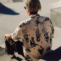 Photo of braided-haired man from behind, sitting on steps above skatepark bowl. He is wearing Blinky Bills Shirt No. 2, a very light pink, over-sized, short-sleeve shirt with a print of different tattoo designs in greyscale.