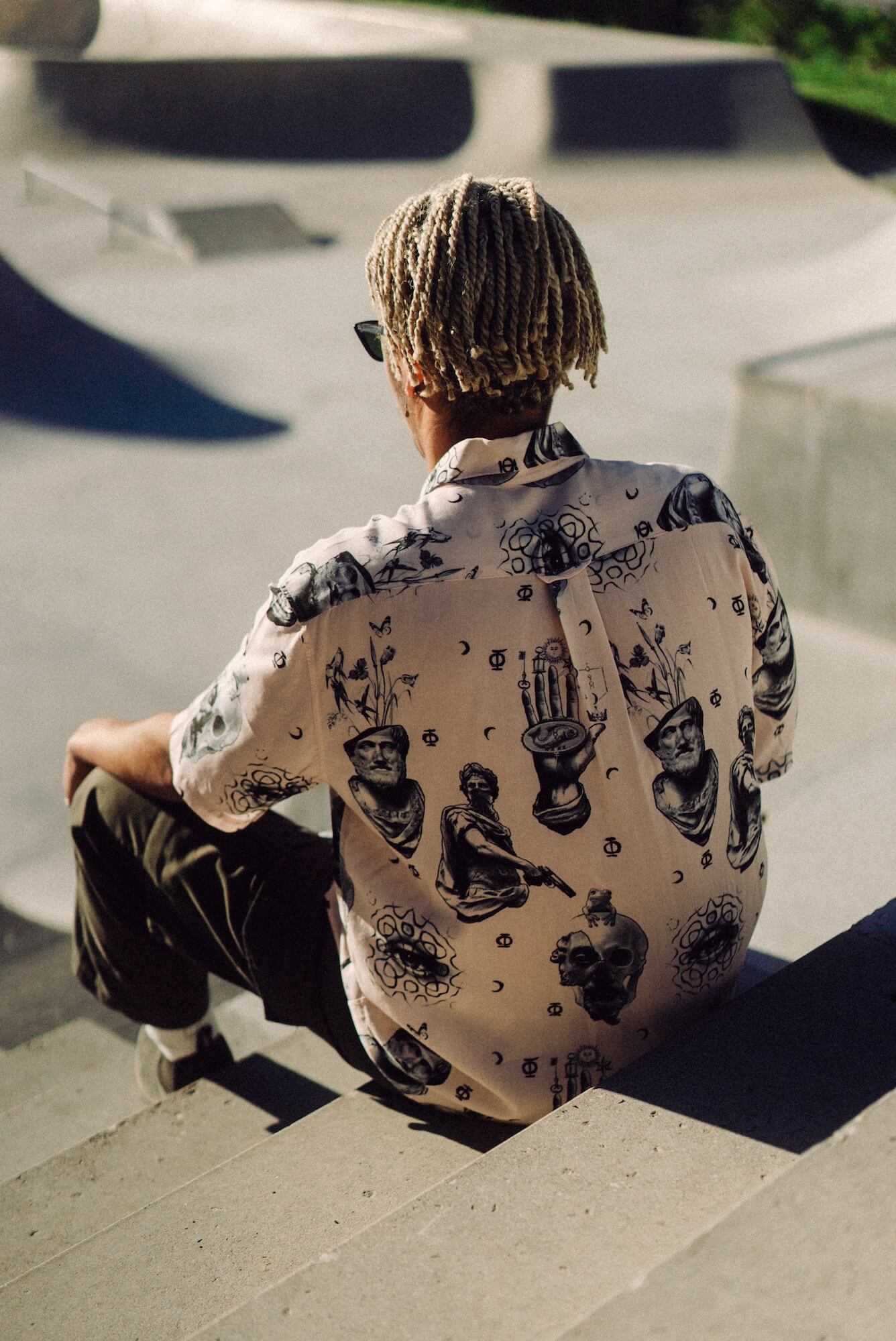 Photo of braided-haired man from behind, sitting on steps above skatepark bowl. He is wearing Blinky Bills Shirt No. 2, a very light pink, over-sized, short-sleeve shirt with a print of different tattoo designs in greyscale.