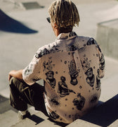 Photo of braided-haired man from behind, sitting on steps above skatepark bowl. He is wearing Blinky Bills Shirt No. 2, a very light pink, over-sized, short-sleeve shirt with a print of different tattoo designs in greyscale.