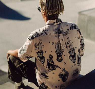 Photo of braided-haired man from behind, sitting on steps above skatepark bowl. He is wearing Blinky Bills Shirt No. 2, a very light pink, over-sized, short-sleeve shirt with a print of different tattoo designs in greyscale.