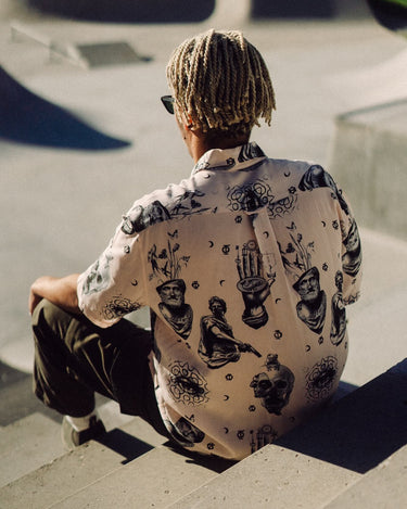 Photo of braided-haired man from behind, sitting on steps above skatepark bowl. He is wearing Blinky Bills Shirt No. 2, a very light pink, over-sized, short-sleeve shirt with a print of different tattoo designs in greyscale.