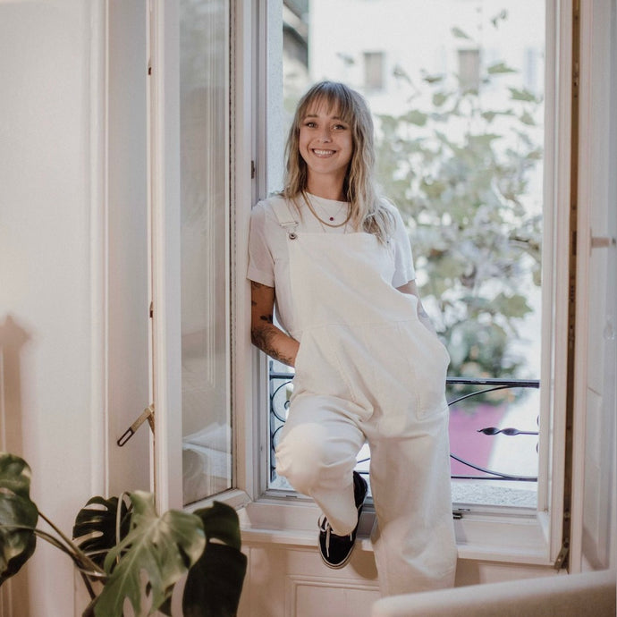 Photo of tattoo artist Kajsa Andersson Rhodiner leaning against white window smiling in white jumpsuit.